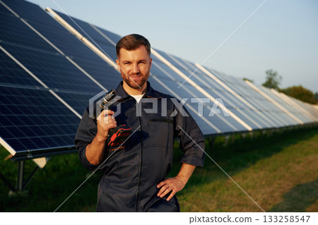 Outside, standing and holding the drill. Man is working with solar panels outdoors at daytime Outside, standing and holding the drill. Man is working with solar panels outdoors at daytime 133258547