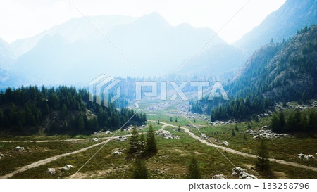 Rolling hills cradle a serene valley, dotted with rocky formations and lush greenery. Paths meander through the landscape, inviting exploration beneath a soft, hazy sky. 133258796