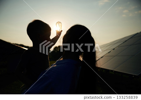 Darkened, against the sun, with light bulb, hope. Mother with her little son are near the solar panels Darkened, against the sun, with light bulb, hope. Mother with her little son are near the solar panels 133258839