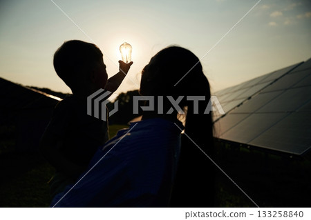 Darkened, against the sun, with light bulb, hope. Mother with her little son are near the solar panels 133258840
