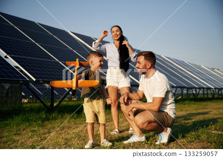 Happy facial expressions, with toy plane. Father, mother and little son are outdoors near solar panels Happy facial expressions, with toy plane. Father, mother and little son are outdoors near solar panels 133259057
