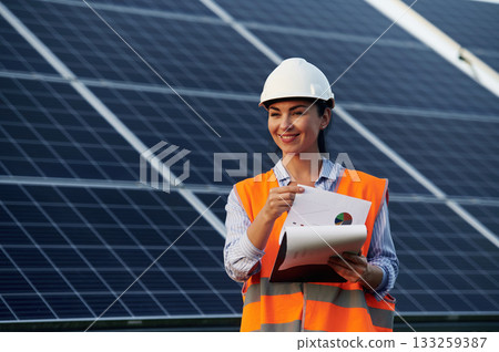 Notepad in hands. Female worker engineer in uniform is near solar panels 133259387