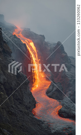 Vertical shot of a bright orange and red river of molten lava cascading down dark, rocky slope of an active volcano. The scene is dramatic and covered in mist or smoke, showcasing geology, raw nature 133260062