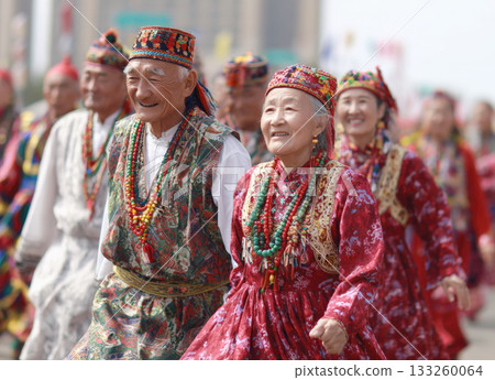 An elderly couple smiling and walking in a parade or cultural festival. They are dressed in vibrant, traditional ethnic costumes with intricate embroidery, colorful beaded jewelry, and unique hats 133260064