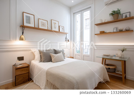 Modern, minimalist bedroom interior with a bright, serene atmosphere. Features a white bed, neutral textiles, wainscoting and crown molding, floating wooden shelves with art, and a glass french door 133260069