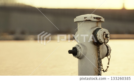 A rusty fire hydrant is positioned by the riverbank, reflecting golden hues of sunset. The water gently glistens in the background, creating a tranquil atmosphere. A rusty fire hydrant is positioned by the riverbank, reflecting golden hues of sunset. The water gently glistens in the background, creating a tranquil atmosphere. 133260385