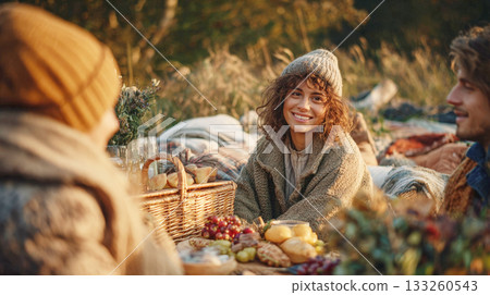 Smiling Young Woman in a Knitted Hat and Cozy Cardigan Enjoying an Autumn Picnic with Friends, Surrounded by Baskets, Food, and Blankets in the Warm Glow of Sunset Smiling Young Woman in a Knitted Hat and Cozy Cardigan Enjoying an Autumn Picnic with Friends, Surrounded by Baskets, Food, and Blankets in the Warm Glow of Sunset 133260543