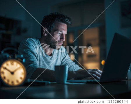 Dedicated Man Working Late at Night on His Laptop in a Dimly Lit Home Office, Illuminated by the Screen Light, Symbolizing Overtime, Concentration, and Remote Work 133260544