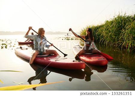 Active weekend, on the water. Women friends are on sup board on the lake together Active weekend, on the water. Women friends are on sup board on the lake together 133261351