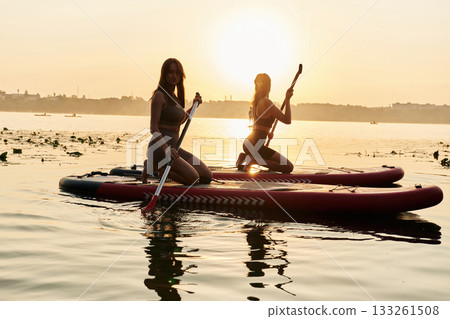 Against sunset light. Women friends are on sup board on the lake together Against sunset light. Women friends are on sup board on the lake together 133261508