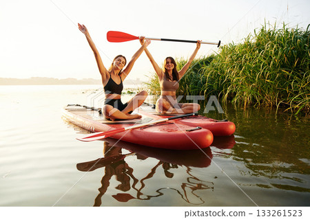 Hands up, smiling and looking forward. Women friends are on sup board on the lake together 133261523