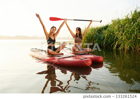 Hands up, smiling and looking forward. Women friends are on sup board on the lake together 133261524