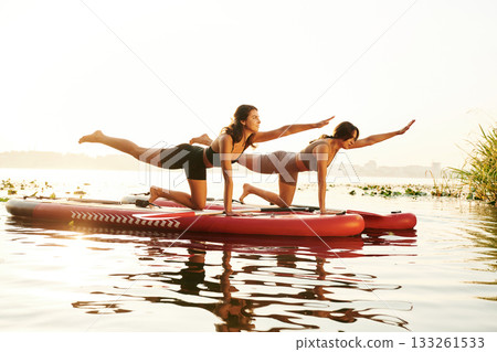 Doing yoga exercises, asana. Women friends are on sup board on the lake together Doing yoga exercises, asana. Women friends are on sup board on the lake together 133261533