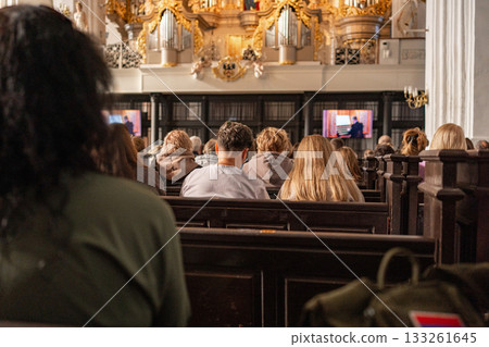 Interior of a historic church with ornate chandeliers and people seated during a service. Warm lighting, wooden benches and classical decor create a spiritual atmosphere Interior of a historic church with ornate chandeliers and people seated during a service. Warm lighting, wooden benches and classical decor create a spiritual atmosphere 133261645