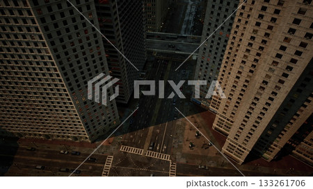 High above a bustling city intersection, towering skyscrapers cast long shadows on the street below. Cars navigate the roads while pedestrians traverse the vibrant urban landscape. 133261706