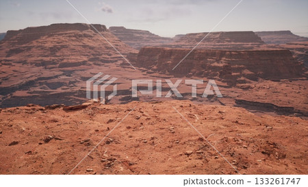 Vast red rock formations extend across the horizon, revealing intricate geological patterns. The sun illuminates the rugged terrain, creating a breathtaking view that draws exploration. 133261747