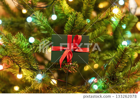 Christmas present on Christmas tree with garland lights , Gift in green box with red ribbon , New Year holidays present 133262078