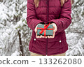 girl faceless in red jacket holding in hands Gift green box with red ribbon against snowy spruce tree in winter forest Christmas and New Year holidays present 133262080