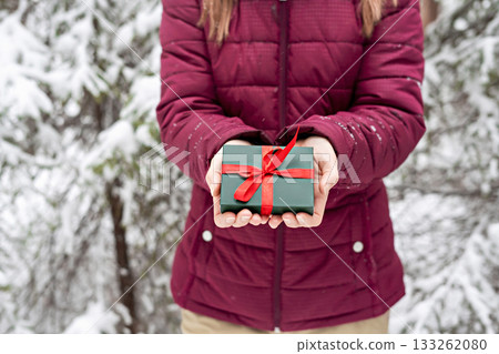 girl faceless in red jacket holding in hands Gift green box with red ribbon against snowy spruce tree in winter forest Christmas and New Year holidays present 133262080