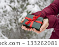 Gift in green box with red ribbon in female hands against snowy spruce tree in winter forest Christmas background and New Year holidays present 133262081