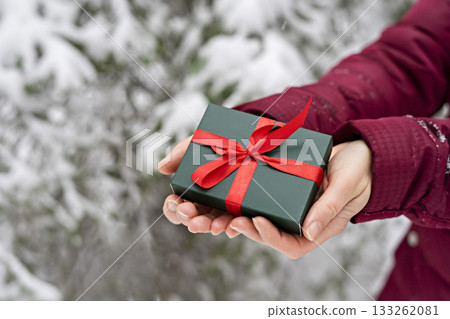 Gift in green box with red ribbon in female hands against snowy spruce tree in winter forest Christmas background and New Year holidays present Gift in green box with red ribbon in female hands against snowy spruce tree in winter forest Christmas background and New Year holidays present 133262081