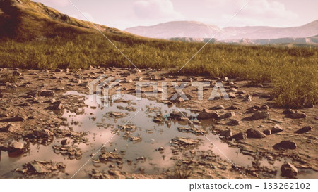 Vibrant hues fill the tranquil landscape, where a small, reflective pool gathers beneath scattered stones. Grassy hills rise in the background, bathed in soft twilight warmth. 133262102