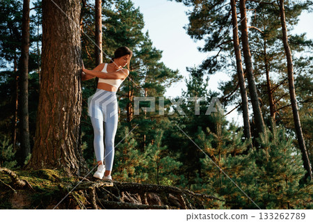 Holding by a tree. Woman in fitness clothes is in the forest Holding by a tree. Woman in fitness clothes is in the forest 133262789