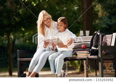 Reading together. Little schoolgirl with her mother are sitting on the bench outdoors 133263092