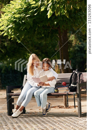Reading together. Little schoolgirl with her mother are sitting on the bench outdoors Reading together. Little schoolgirl with her mother are sitting on the bench outdoors 133263096