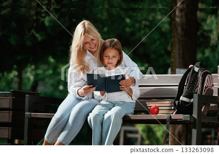 Information in notepad, reading it. Little schoolgirl with her mother are sitting on the bench outdoors Information in notepad, reading it. Little schoolgirl with her mother are sitting on the bench outdoors 133263098