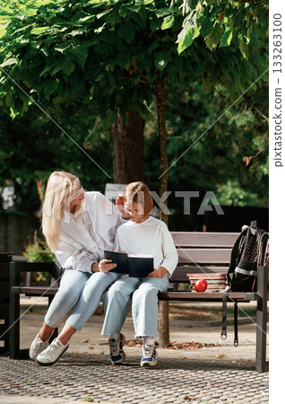 Information in notepad, reading it. Little schoolgirl with her mother are sitting on the bench outdoors Information in notepad, reading it. Little schoolgirl with her mother are sitting on the bench outdoors 133263100