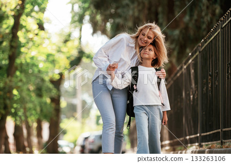 Front view, walking. Schoolgirl with her mother are outdoors together Front view, walking. Schoolgirl with her mother are outdoors together 133263106