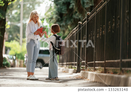 With books, front view. Schoolgirl with her mother are outdoors together With books, front view. Schoolgirl with her mother are outdoors together 133263148