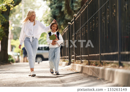 With books, front view. Schoolgirl with her mother are outdoors together With books, front view. Schoolgirl with her mother are outdoors together 133263149