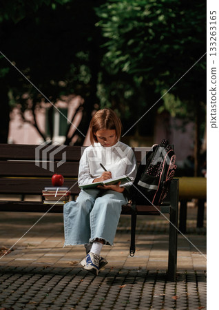 Writing ideas into notepad. Schoolgirl is sitting on the bench with a bunch of books outdoors Writing ideas into notepad. Schoolgirl is sitting on the bench with a bunch of books outdoors 133263165