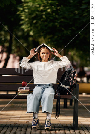 Having fun, book over the head. Schoolgirl is sitting on the bench with a bunch of books outdoors Having fun, book over the head. Schoolgirl is sitting on the bench with a bunch of books outdoors 133263170