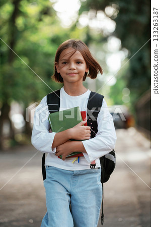 Walking forward and holding notepad. Schoolgirl with backpack is outdoors Walking forward and holding notepad. Schoolgirl with backpack is outdoors 133263187