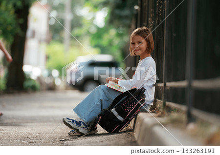Sitting on the sidewalk. Schoolgirl with backpack is outdoors Sitting on the sidewalk. Schoolgirl with backpack is outdoors 133263191