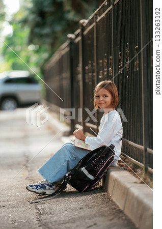 Sitting on the sidewalk. Schoolgirl with backpack is outdoors Sitting on the sidewalk. Schoolgirl with backpack is outdoors 133263192