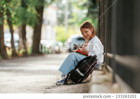 Smartphone in hand, sitting. Schoolgirl with backpack is outdoors 133263201