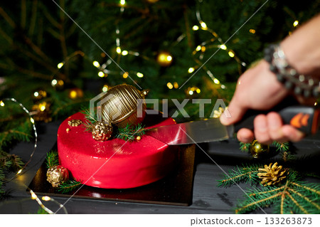 A Person Cuts a Festive Red Cake Adorned With Decorative Ornaments and Greenery During a Joyful Holiday Celebration in a Cozy Setting 133263873