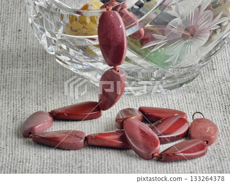 Natural stone, irregularly shaped red jasper beads in crystal basket with stones. Burlap background Natural stone, irregularly shaped red jasper beads in crystal basket with stones. Burlap background 133264378