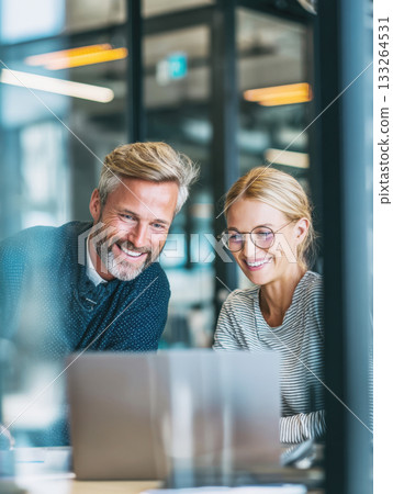 Happy, smiling business colleagues working together on a laptop in a modern glass office. The image captures successful collaboration, positive teamwork, and a contemporary corporate environment Happy, smiling business colleagues working together on a laptop in a modern glass office. The image captures successful collaboration, positive teamwork, and a contemporary corporate environment 133264531