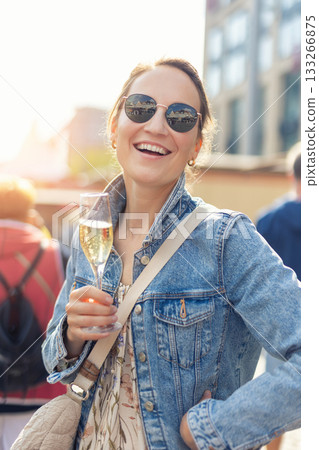 Portrait young adult pretty happy woman woman hold sparkling wine prosecco drink in lively street setting. Denim jacket floral dress sunglasses create vibrant atmosphere filled with festive energy Portrait young adult pretty happy woman woman hold sparkling wine prosecco drink in lively street setting. Denim jacket floral dress sunglasses create vibrant atmosphere filled with festive energy 133266875