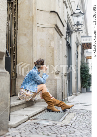 Portrait of young adult woman wear floral dress denim jacket brown suede boots sitting on stairs outdoors in old European city. Stylish female person enjoy walk travel urban lifestyle summertime day Portrait of young adult woman wear floral dress denim jacket brown suede boots sitting on stairs outdoors in old European city. Stylish female person enjoy walk travel urban lifestyle summertime day 133266985