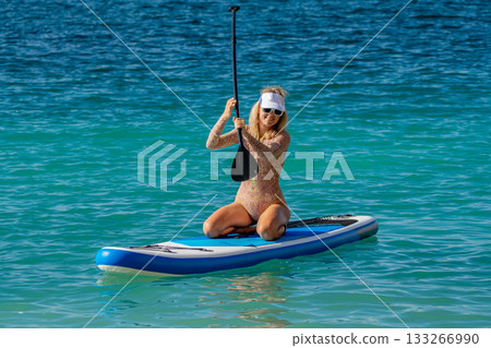 Paddleboarding, woman, ocean. Smiling woman kneeling on a stand-up paddleboard in clear blue water, enjoying summer recreation with copy space Paddleboarding, woman, ocean. Smiling woman kneeling on a stand-up paddleboard in clear blue water, enjoying summer recreation with copy space 133266990