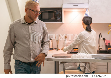 Here's a title attempting to follow all instructions: Couple Kitchen Birthday Surprise; Man entering; Woman cooking; celebratory mood, modern apartment. Here's a title attempting to follow all instructions: Couple Kitchen Birthday Surprise; Man entering; Woman cooking; celebratory mood, modern apartment. 133267042