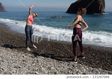 Women fitness stretching partners warm up on a sunny pebble beach by the ocean Women fitness stretching partners warm up on a sunny pebble beach by the ocean 133267066