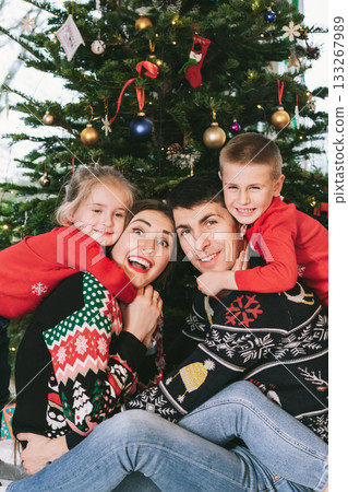 Smiling family portrait in festive sweaters sitting with gifts under Christmas tree at home during holiday. Happy parents and kids cuddling, feeling excited. Family time together. Happy childhood 133267989