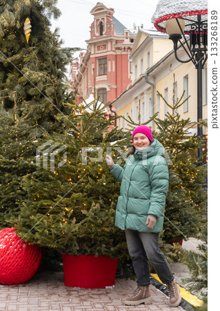 A woman in a green winter coat stands among decorated Christmas trees with warm yellow lights on a snowy city street, creating a festive atmosphere A woman in a green winter coat stands among decorated Christmas trees with warm yellow lights on a snowy city street, creating a festive atmosphere 133268189